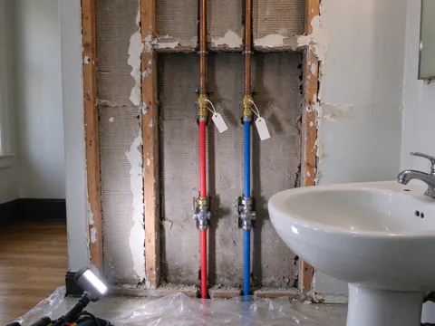 New red and blue PEX lines joined to older copper at tagged brass transition fittings inside an opened bathroom wall in a San Francisco flat